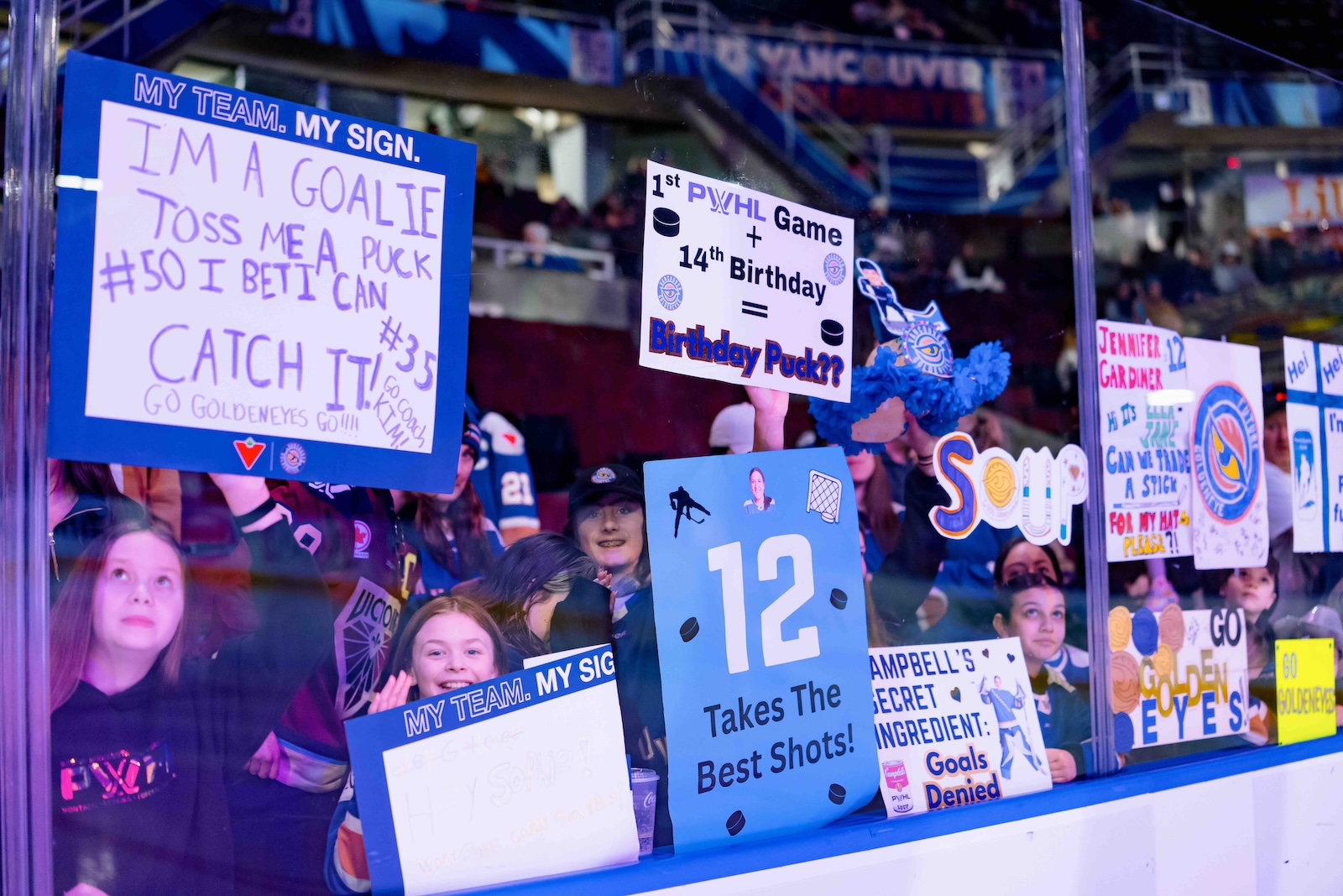 Top: A female hockey player standing on the ice talks to fans through the glass. Bottom: Young girls at a hockey game press signs up against the glass. The signs read: &ldquo;I&rsquo;m a goalie, pass me a puck I bet I can catch it!&rdquo; and &ldquo;1st PWHL game + 14th birthday = birthday puck?&rdquo;