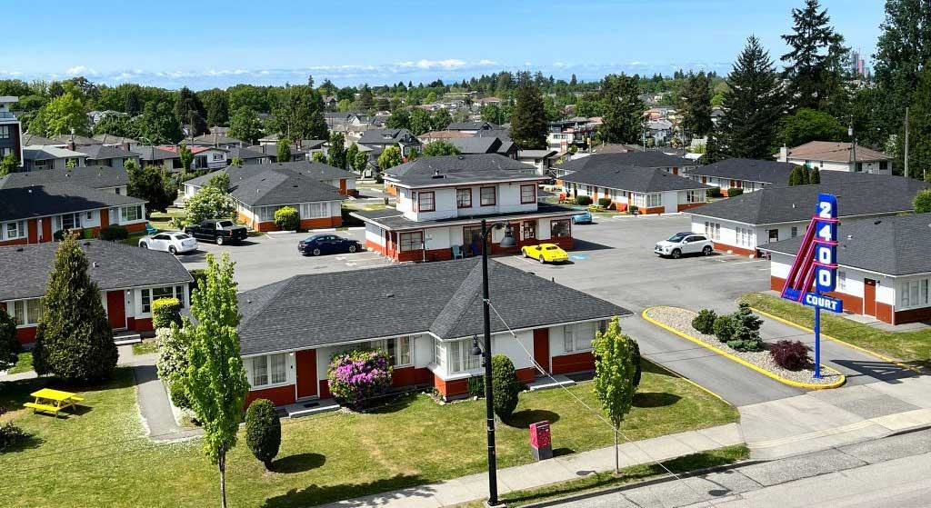 A panorama view of the 2400 Motel, a series of short grey-roofed buildings with white stucco and orange trim across a sprawling concrete lot on a sunny day. To the right of the frame is a blue and red neon sign that reads &ldquo;2400 Court&rdquo; in white letters.