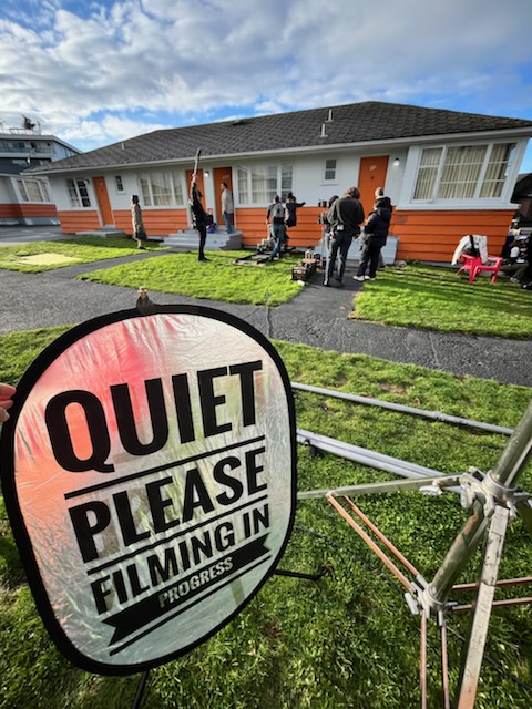 In the foreground, a silver sign with black text reads &ldquo;Quiet please. Filming in progress.&rdquo; Behind the sign is a film crew on the steps of a one-storey bungalow with orange trim, stucco exterior walls and grey roofing.