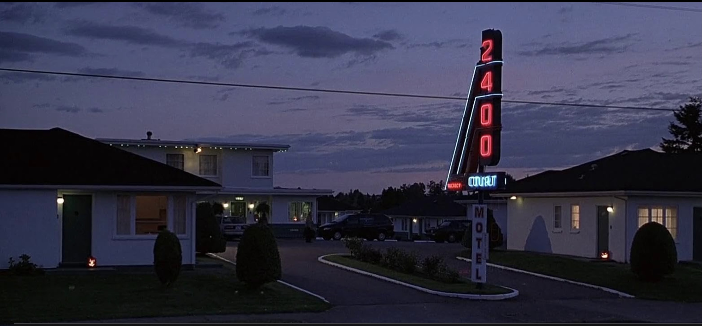 A nighttime landscape photograph of the 2400 Motel, which features several one-storey white bungalows against a darkening sky. In the centre of the frame is a neon sign with red and blue letters that reads &ldquo;2400 Court.&rdquo;