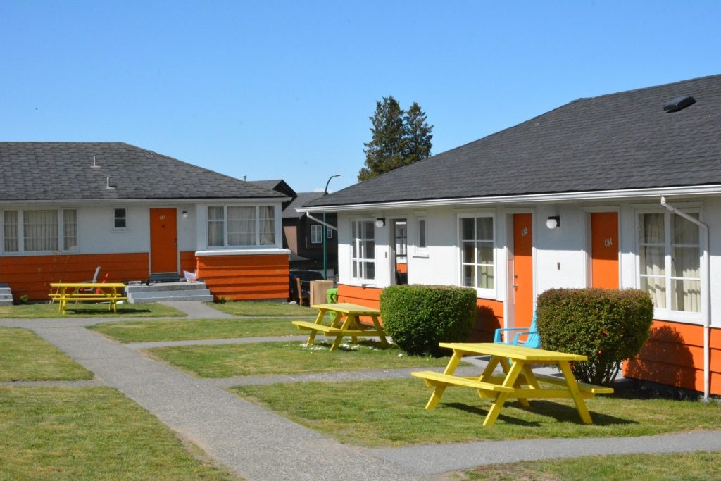 Two one-storey bungalows have white walls, orange trim, grey roofs and yellow picnic tables on their front lawns. Above them is a bright blue sky.
