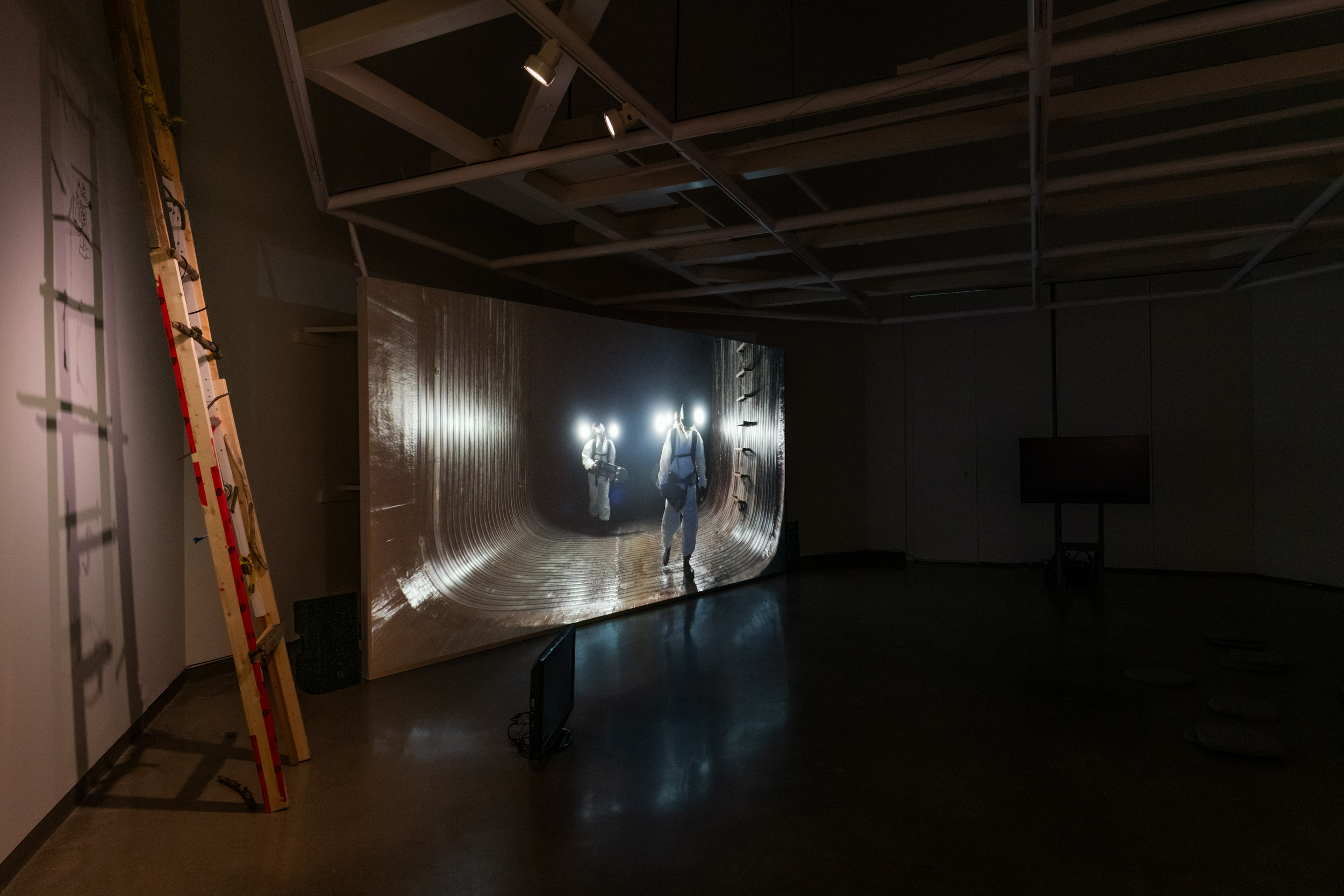 A photograph of a large-format video installation in a large darkened room. A floor-to-ceiling screen depicts two figures in white Hazmat suits with helmets and headlamps walking through a tunnel. To the left of the frame, a ladder leans against a wall.  