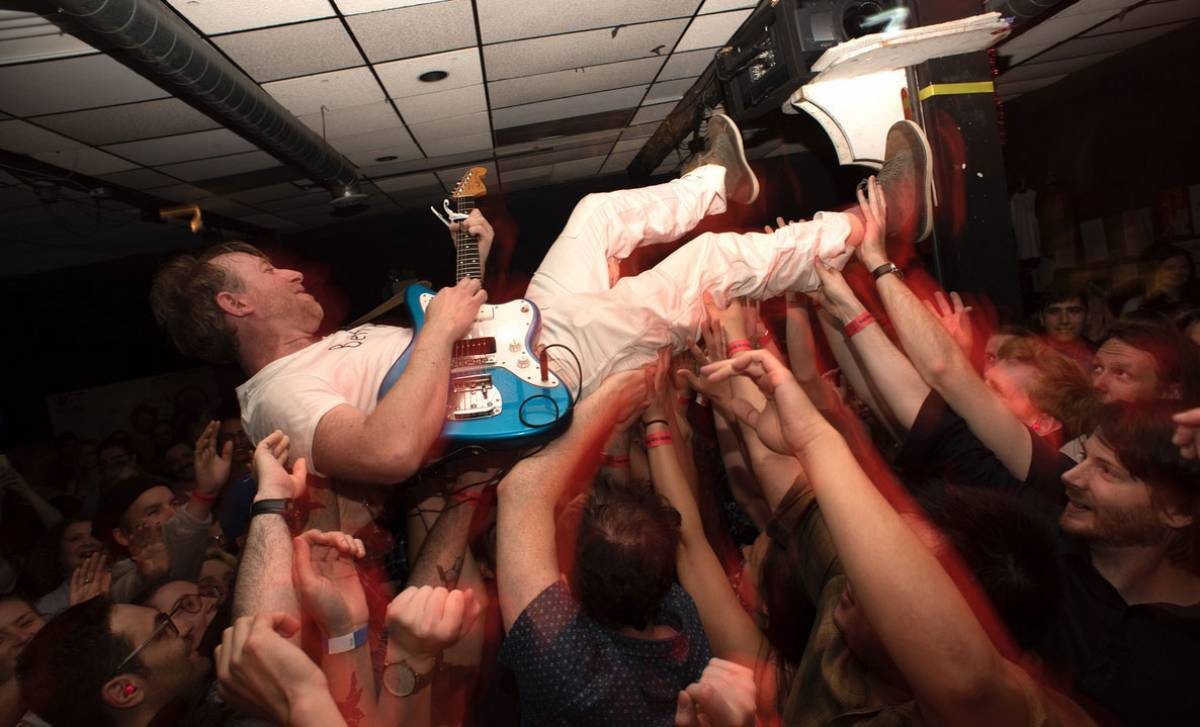 A performer wearing a white T-shirt and white trousers crowd-surfs in a small indoor venue. He is holding and playing a light blue electric guitar.  