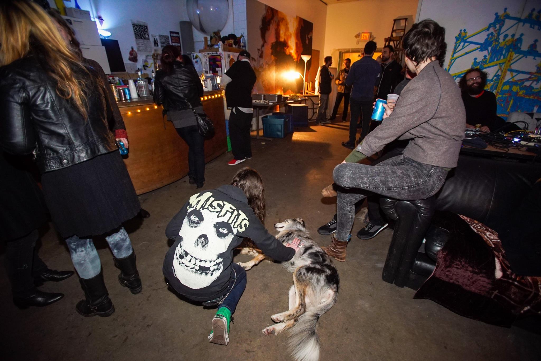 Two stacked images depict, at top, an audience watching, at close range, while a rock band plays to the left of the frame; and at bottom, people lounging in an indoor venue space. A person in a Misfits hoodie is petting a dog lying on the floor.