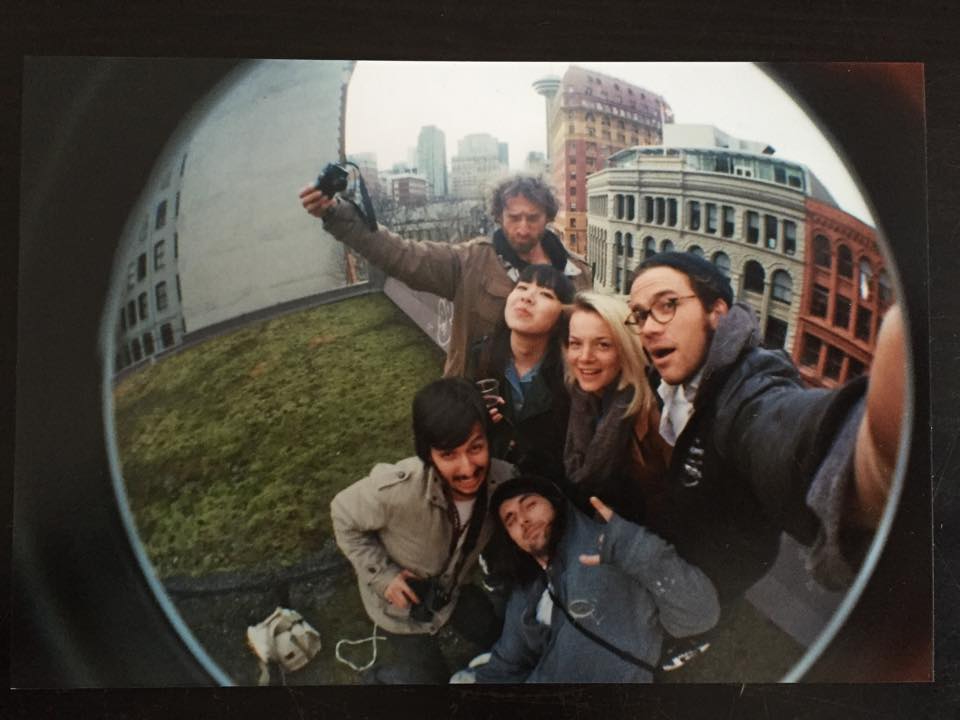 A vintage-looking photograph taken with a fisheye lens depicts a group of six people posing in a grassy area on a downtown rooftop on a grey day.