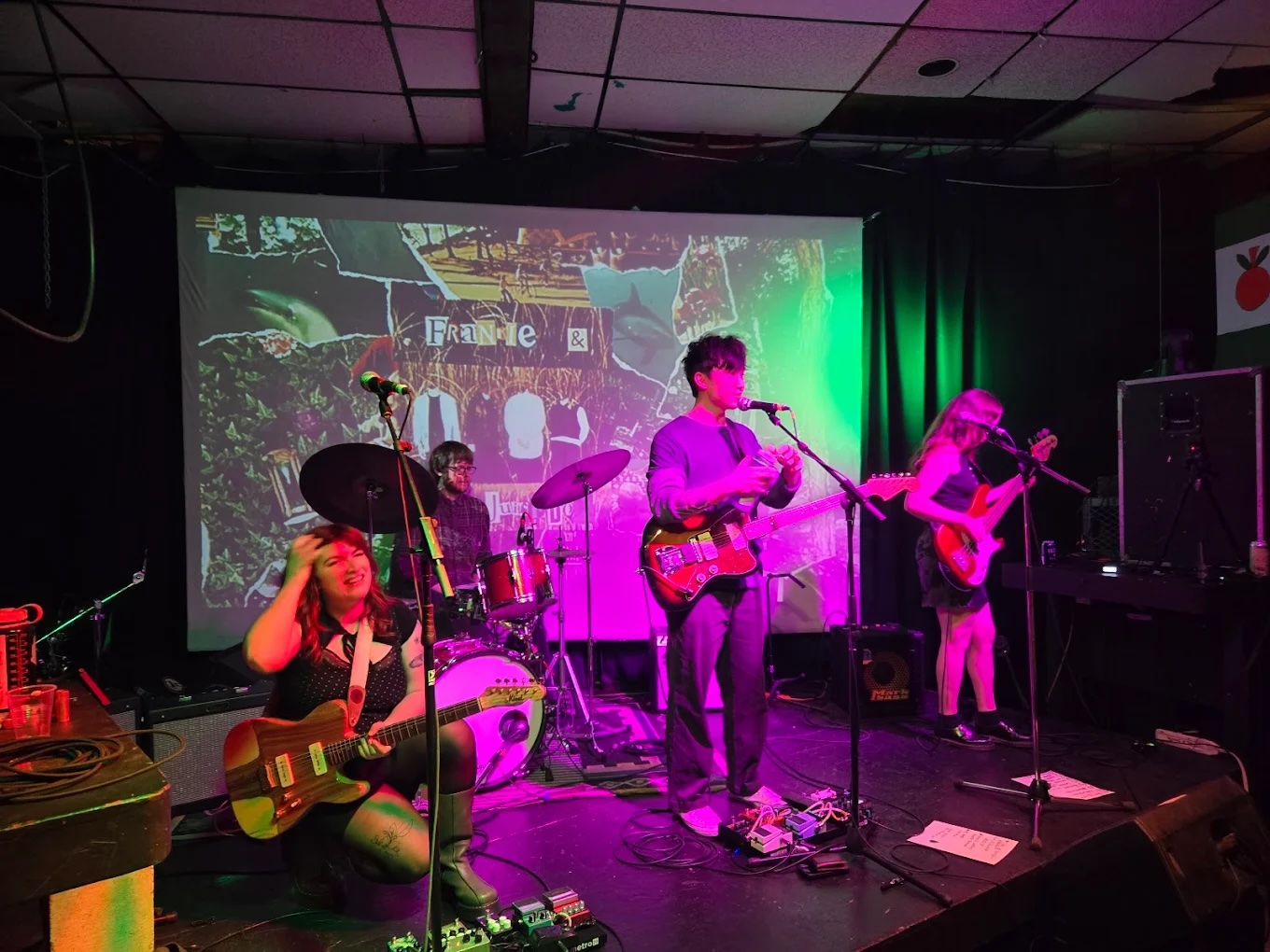 A rock band pauses between songs on a small black stage. Its young members are lit with colourful lights. A projected screen that reads &ldquo;Frankie & the Juicebox&rsquo; is behind them.
