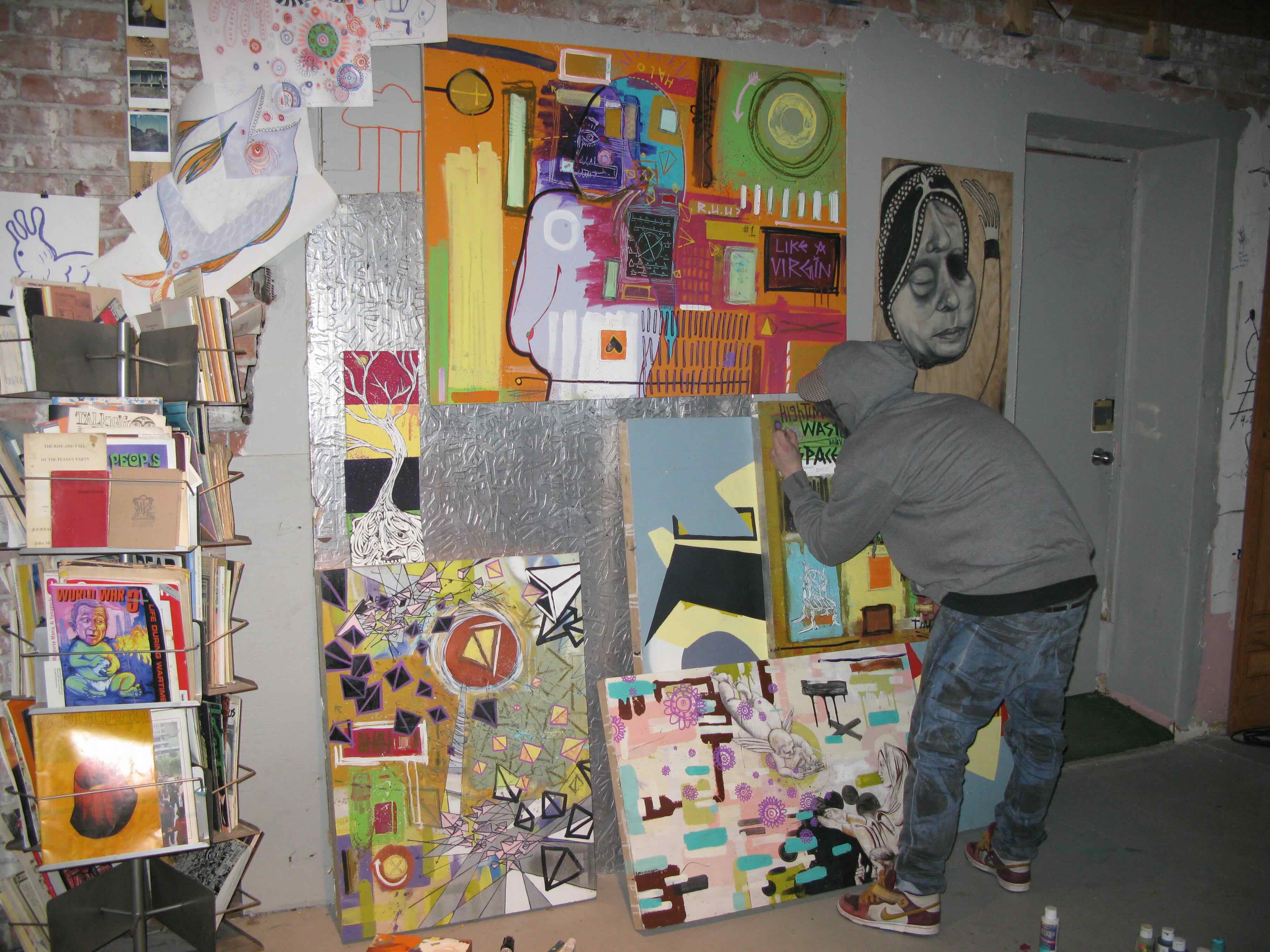 An artist in a grey hoodie and jeans has his back turned to the camera. He is tinkering with one of several colourful paintings arranged on the wall in a small indoor space.