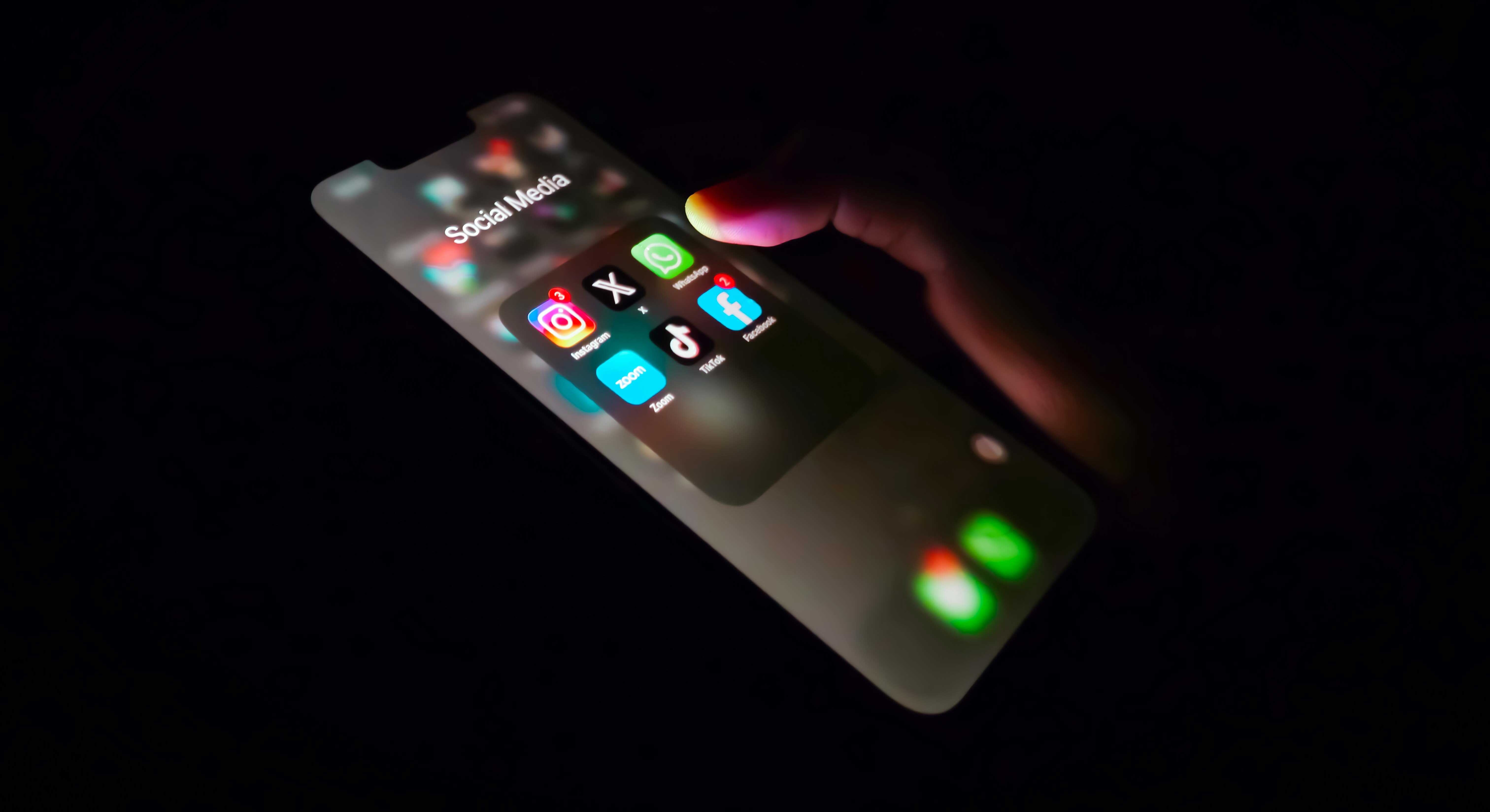 A hand holds a smartphone in the dark, poised over a menu of social media apps including Instagram, X, WhatsApp, Facebook and Threads.
