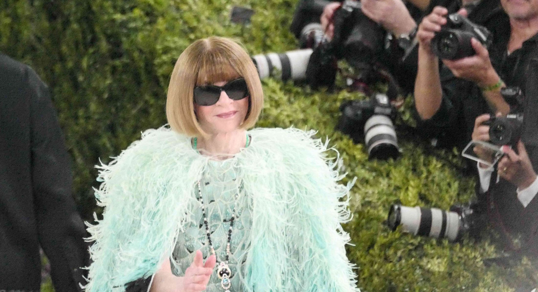 Anna Wintour has short dark hair and sunglasses. She is wearing a feathered light turquoise jacket and raising her hand to wave. People with cameras are lined up against a hedge to the right of the frame.