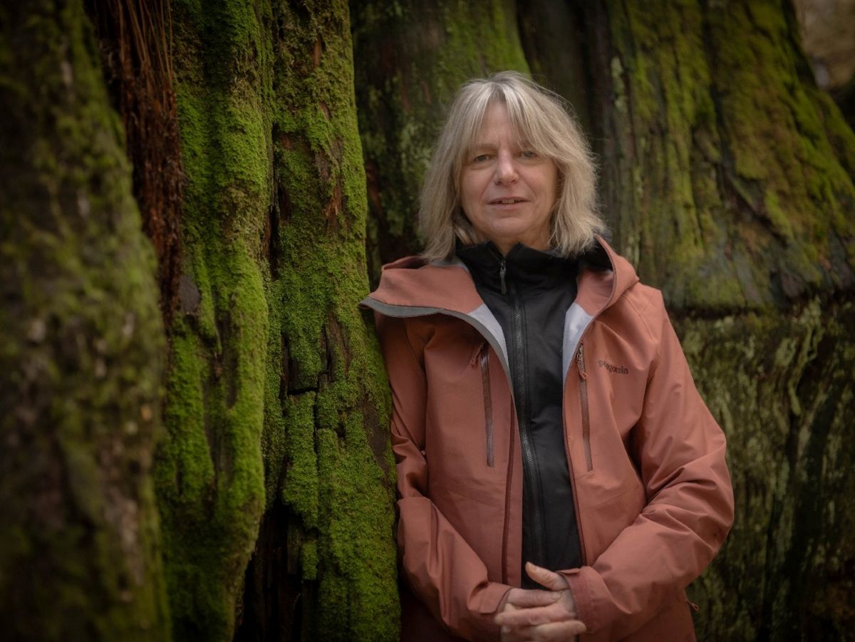 Suzanne Simard stands to the right of the frame. She has wavy blond hair cut just above her shoulders. She is wearing a pink windbreaker over a black waterproof jacket while standing against a large tree trunk covered in thin green moss.