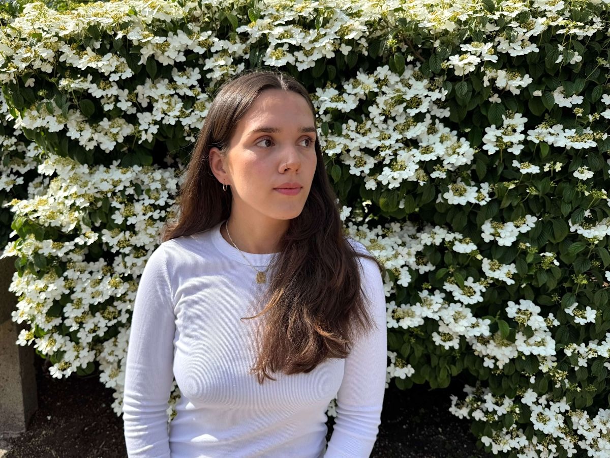 Anjini Snape, a woman with light skin and long brown hair, stands in front of a white-flowering bush, looking toward the right of the frame.