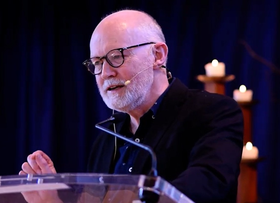 A bald man with a grey-white beard and glasses speaks at a lectern. Candles are visible behind him.