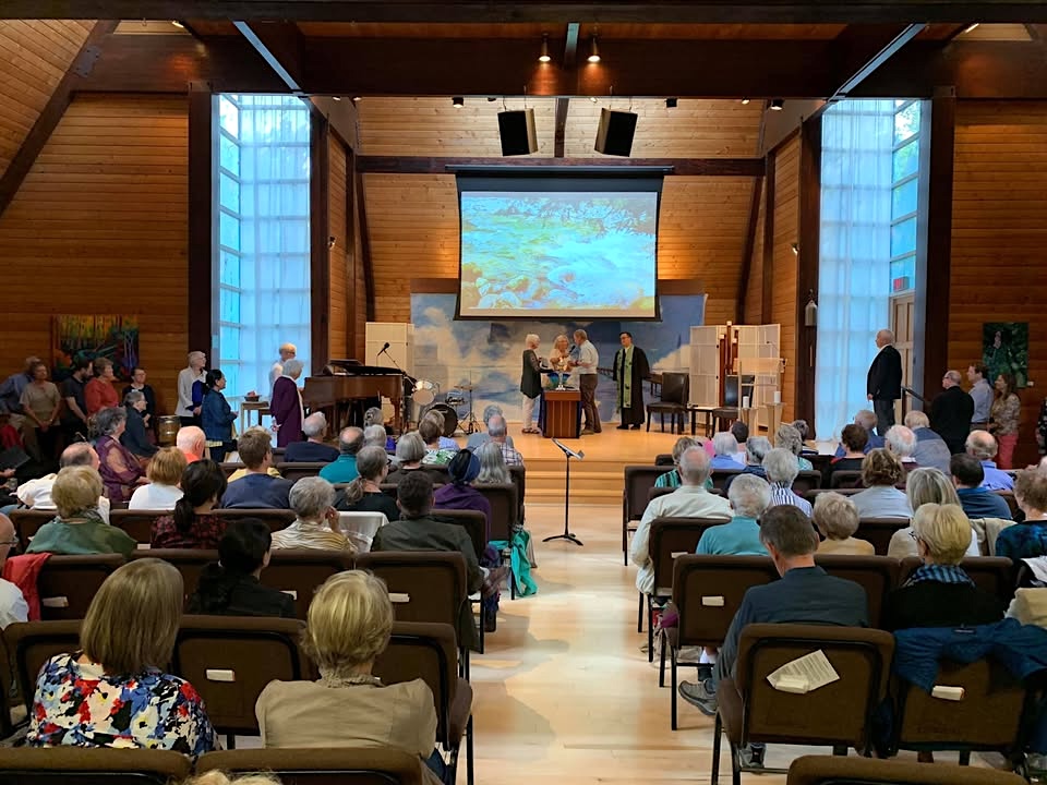 People sit in rows of seats at a service inside a wood-panelled church hall. 