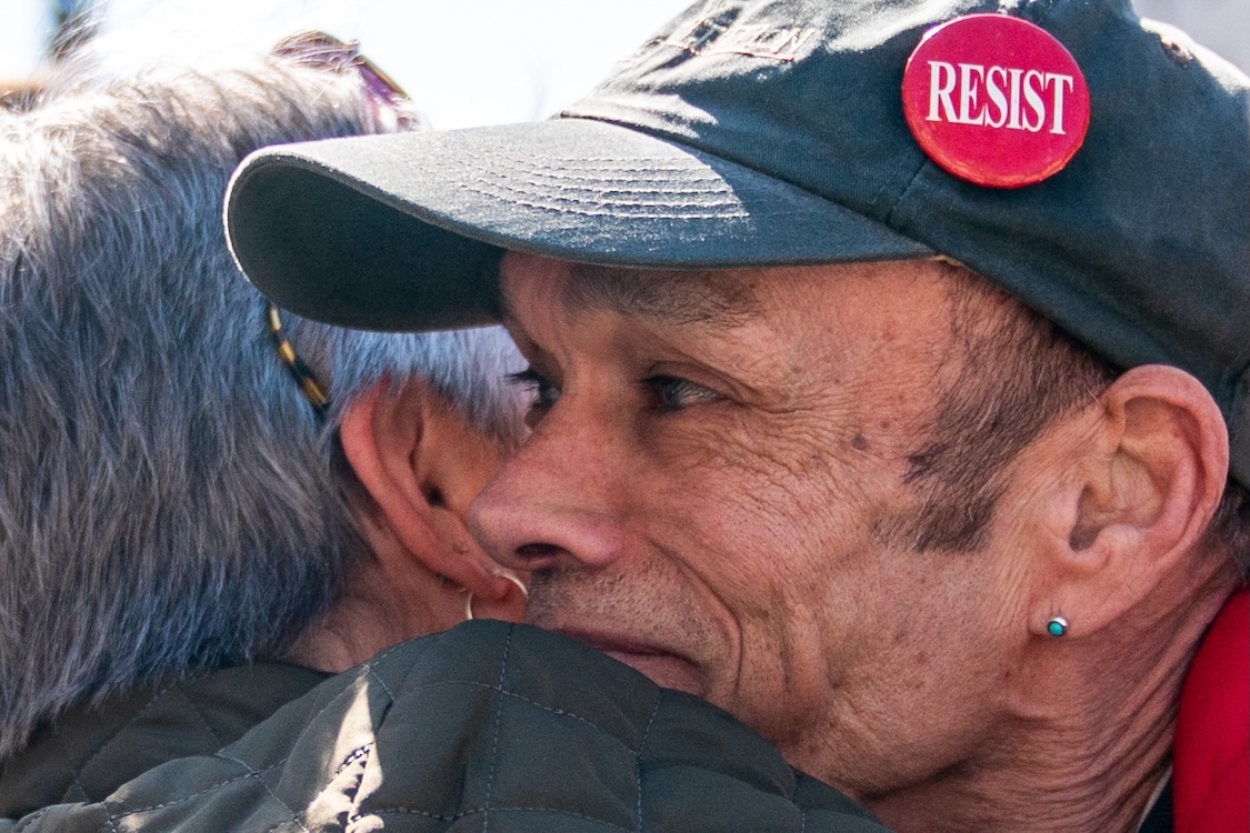A close-up photo of two people in emotional embrace. The person on the left has their back turned to the camera. They have grey hair and earrings. The person on the right is photographed in profile; they have dark hair and are wearing a turquoise earring and a grey cap with a red button that reads &ldquo;RESIST.&rdquo;