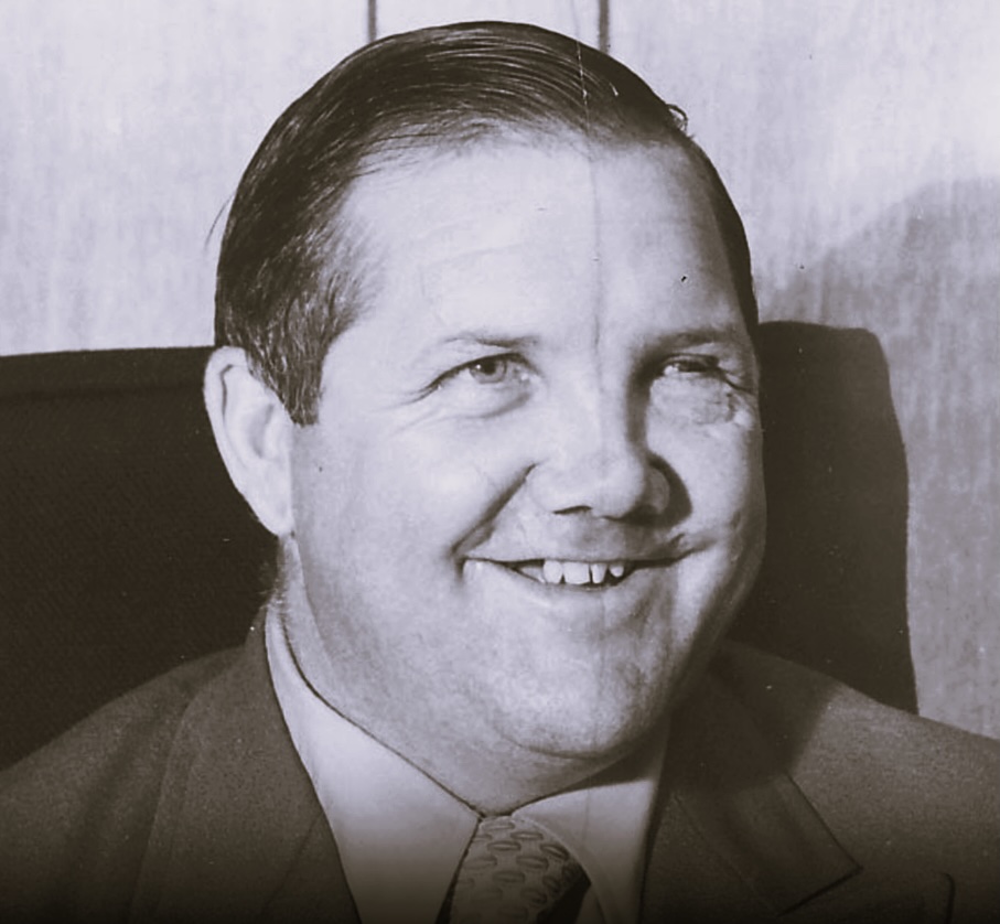 A round-faced, light-skinned man with slicked-back dark hair smiles broadly at the camera.