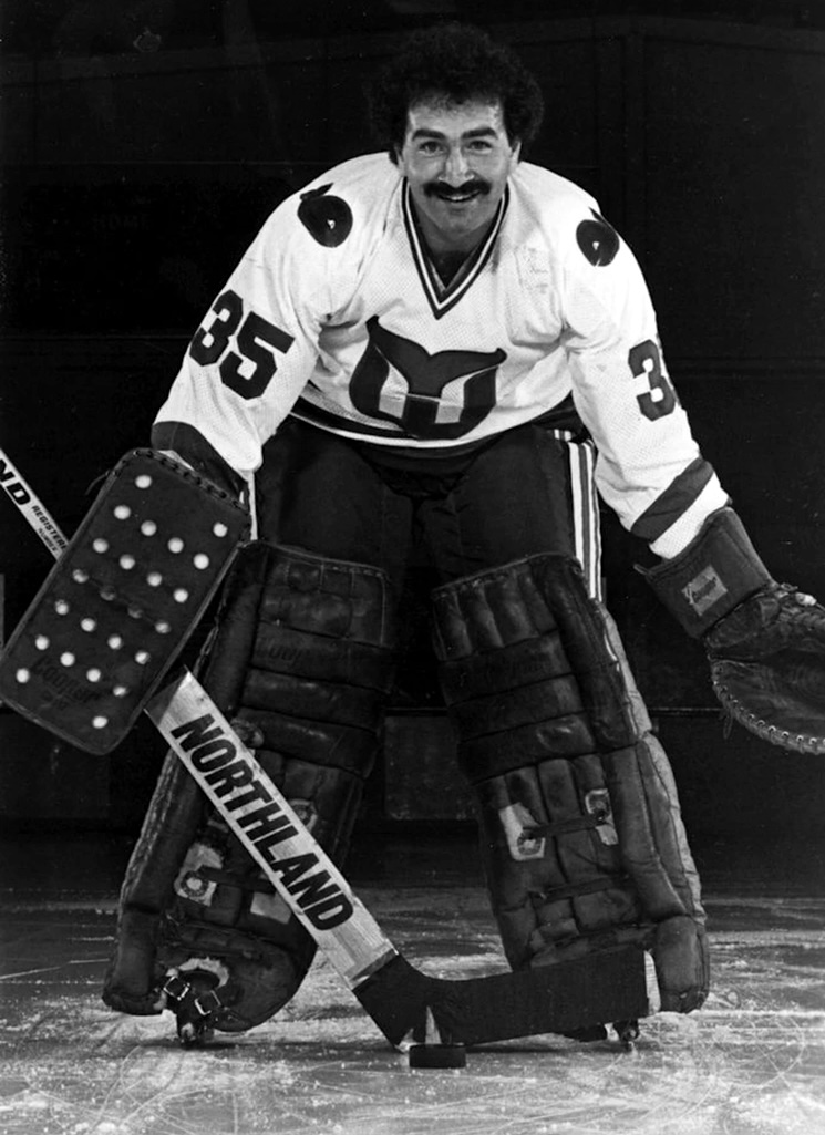 A dark-haired man with a moustache poses in a white jersey and goalie pads.
