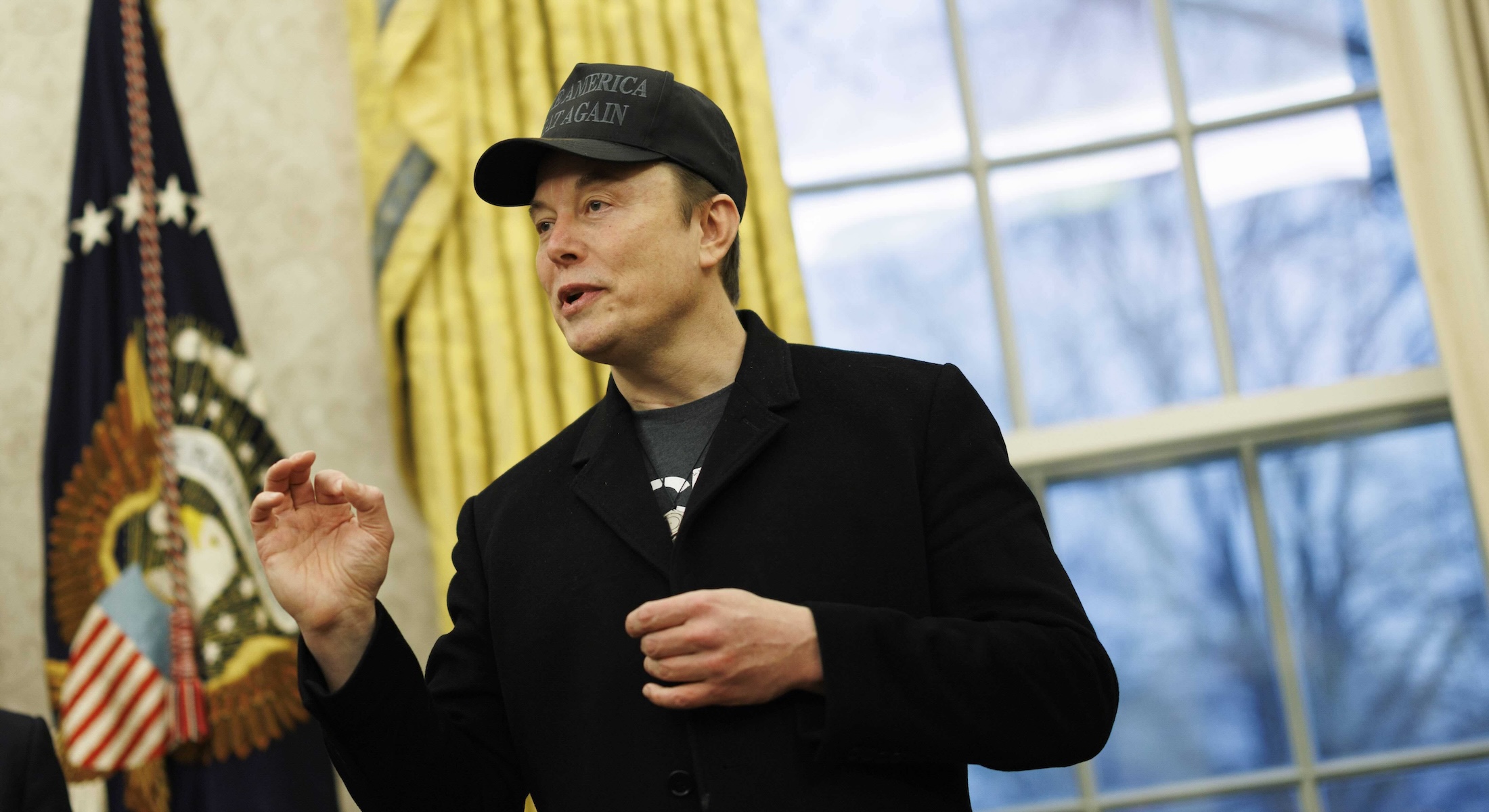 Elon Musk wears a black ball cap and black blazer over a black T-shirt. He is standing in the Oval Office, mid-speech and gesticulating with his right hand. Behind him are gold curtains and an American flag.