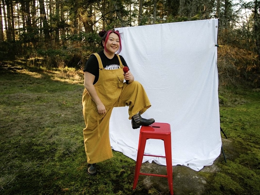 Scarlet Chen is wearing oversized yellow overalls over a black T-shirt. She has long black hair tied up in two buns and dyed fuchsia strands framing her face. She holds a fuchsia microphone with a leg up on a red stool against a white drop sheet. She is standing in the grass with a stand of sun-dappled trees in the background.