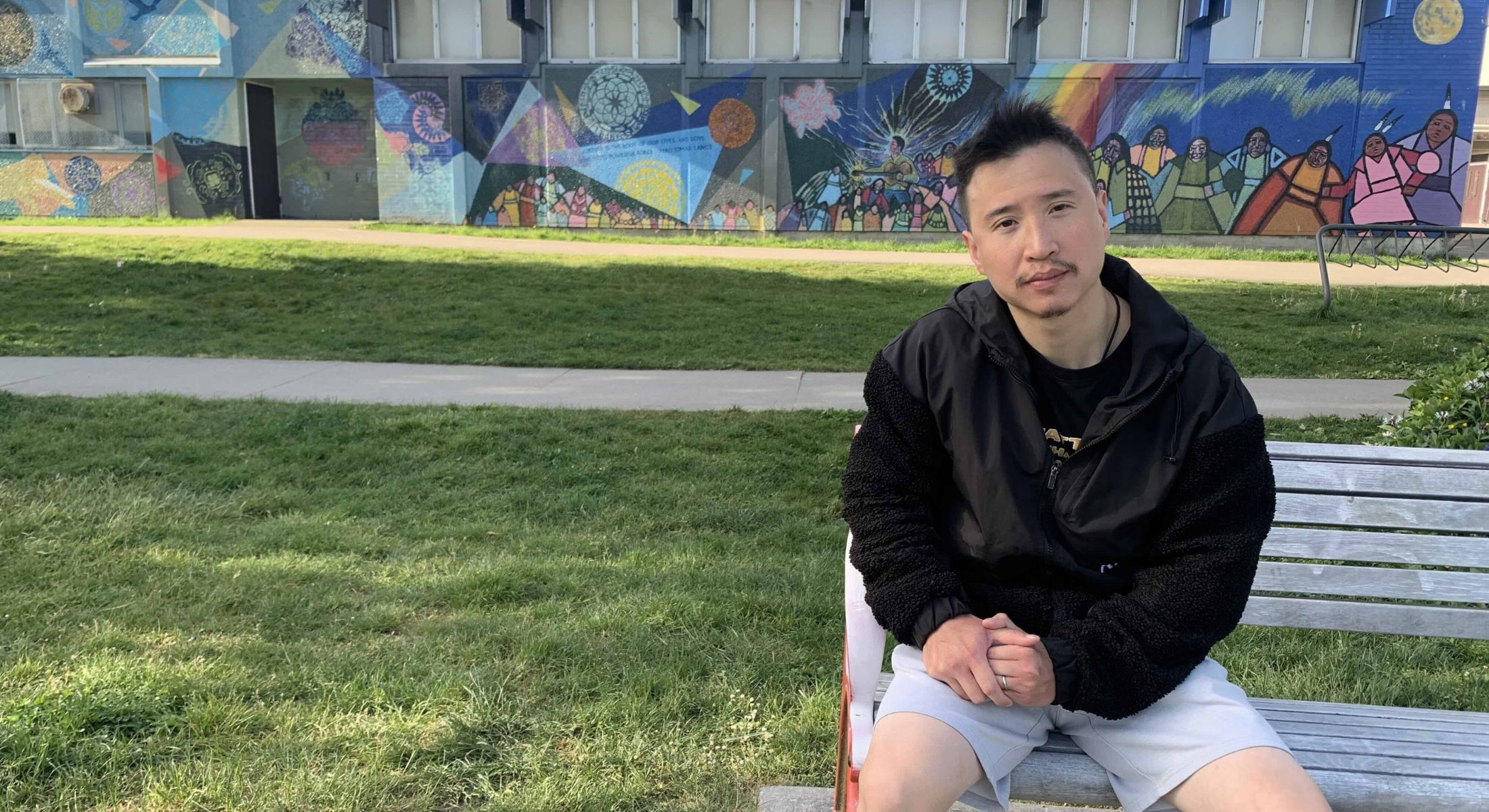 Steven Hsu is seated on a bench outdoors in a grassy area. Behind him is a sidewalk and a school wall featuring a colourful mural. Hsu has short spiky black hair and he is wearing a black hooded jacket and grey gym shorts.