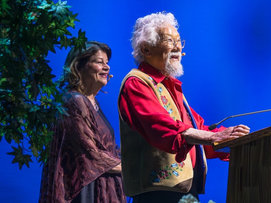 David Suzuki, with white hair and goatee, wearing a red collared shirt under an embroidered vest, speaks at a lectern.