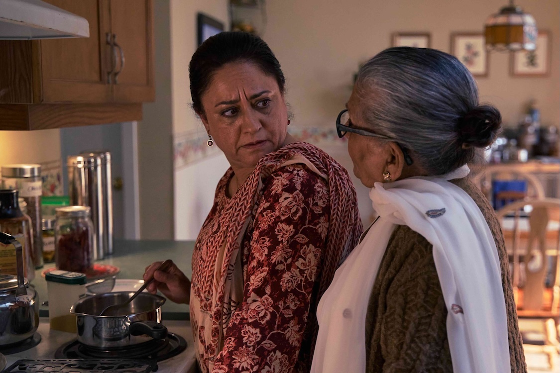 Two actors interact in a still from a TV series. They are standing in the kitchen of a house. On the left, actor Sandeep Bali stands stirring a small pot on a white electric stove. She has dark hair pulled back and she is wearing a sari with a raspberry floral pattern. She turns quizzically to actor Usha Kiran Uppal, right, who has greying hair pulled back in a bun, glasses and a white sari under a taupe wool cardigan. 