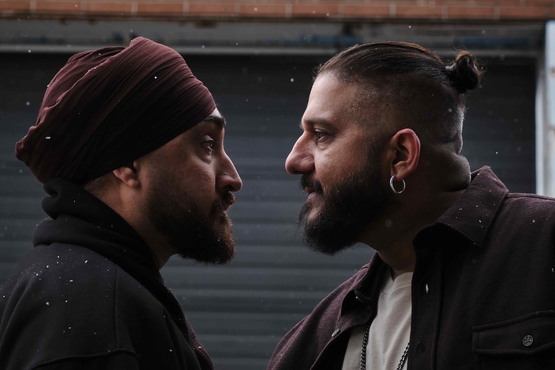 Actors Jasmeet Raina, left, and Amit Aujila, right, stand facing each other in a confrontation. Raina has a beard and is wearing a burgundy turban. Aujila has a beard, dark hair tied in a bun, and a silver hoop earring. It&rsquo;s snowing lightly and a grey garage door is in the background.