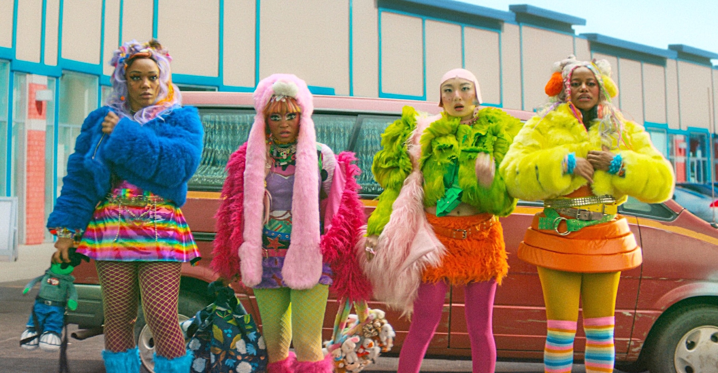 From left, actors Naomi Ackie, Taylour Paige, Poppy Liu and Keke Palmer stand in a row in front of a beat-up red minivan in an outdoor parking lot in a movie scene. They are all wearing rainbow-coloured, furry outfits and over-the-top accessories. 