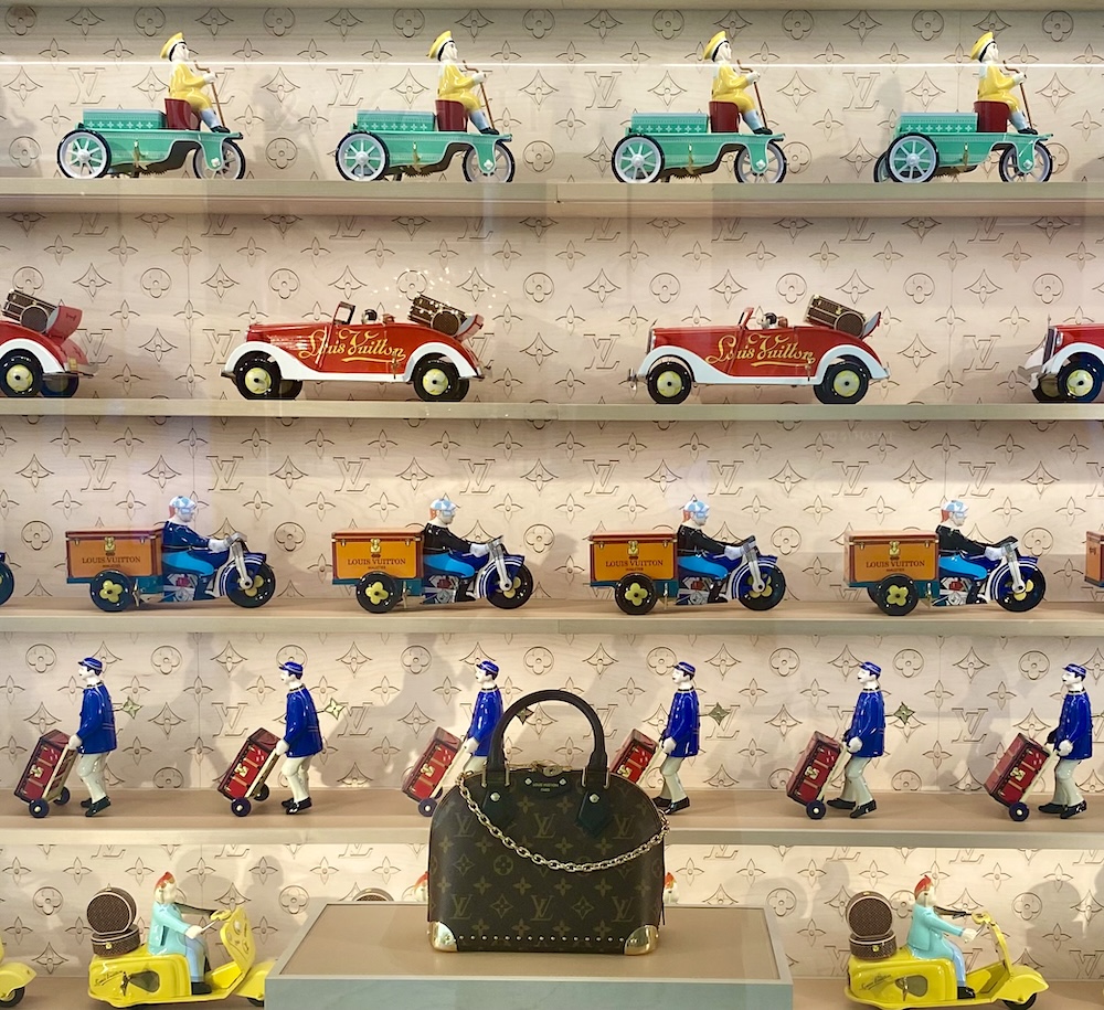 A small brown leather handbag embossed with the Louis Vuitton logo sits on display against a backdrop of rows of ceramic antique-style Louis Vuitton figurines depicting old cars, delivery workers and motorcyclists against a wallpaper of Louis Vuitton logos.