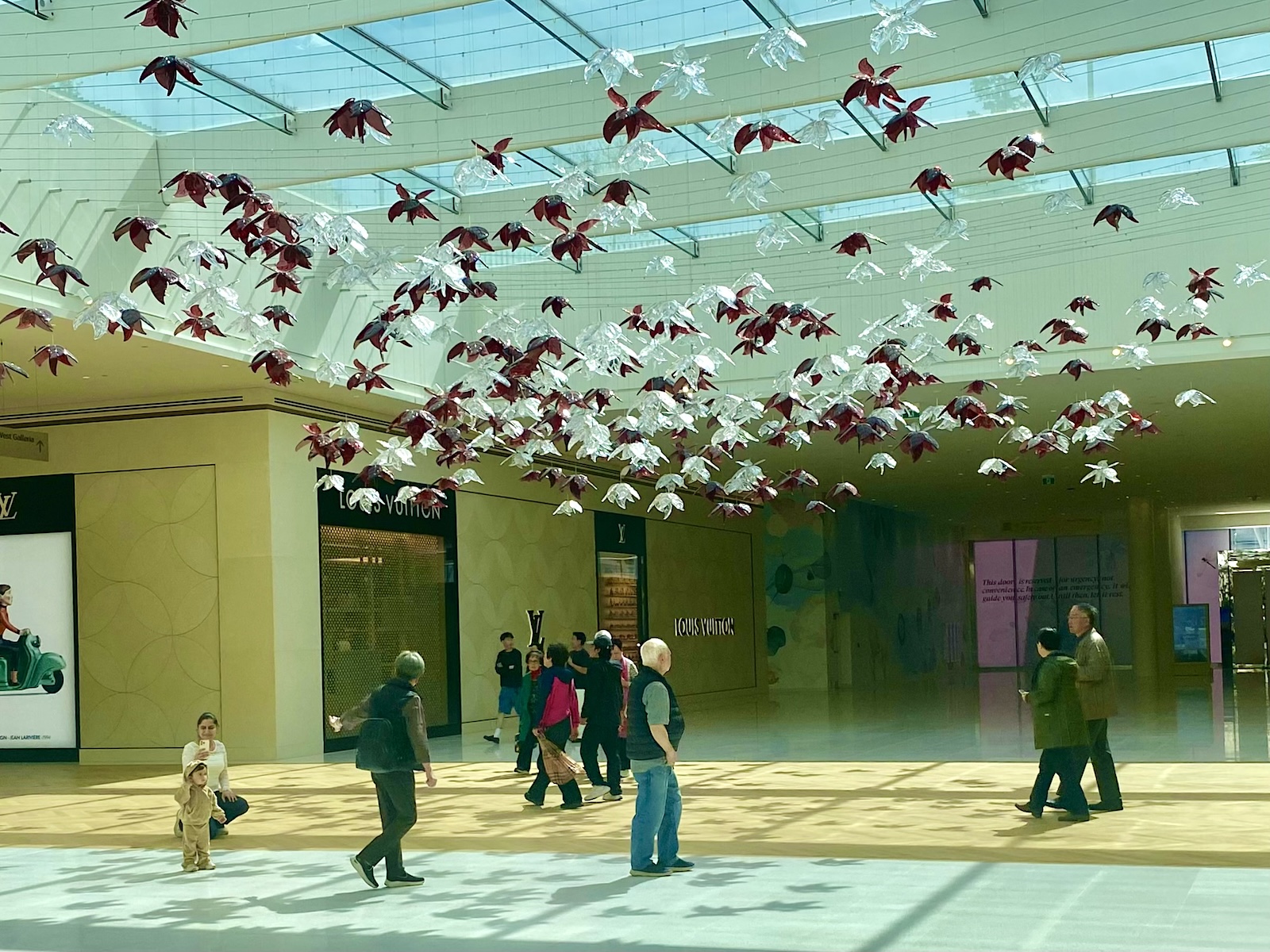 People walk in a hallway of an indoor shopping mall. Hanging from the ceiling is a red and white art installation of floral shapes.