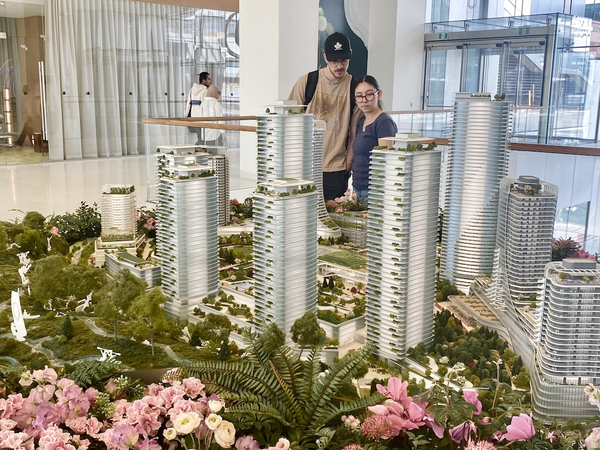 A young man and woman wearing glasses and casual streetwear look at an architectural model of a new development with several residential towers and a green space against a foreground of artificial pink flowers and greenery.