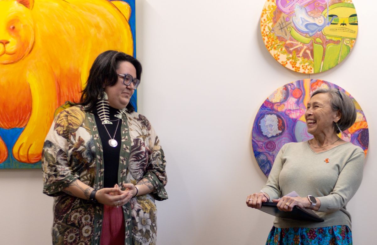Two women stand together in a gallery space where colourful artwork is on the white walls. Shay Paul, left, has wavy dark shoulder-length hair and glasses. She is wearing fish skeleton earrings, a floral top over a black turtleneck, and looking at Elder Joanne Brown, right, and smiling. Brown, right, is slightly shorter than Paul. She has short light brown hair and she is laughing. She wears a taupe cotton shirt with a small pin bearing an orange T-shirt. She is wearing a bright blue floral skirt.