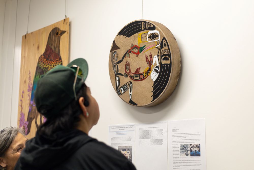 A person wearing a green ball cap stands with their back angled towards the camera, looking at a painted drum hanging on a white wall. They have short dark hair and they are wearing a dark hoodie.