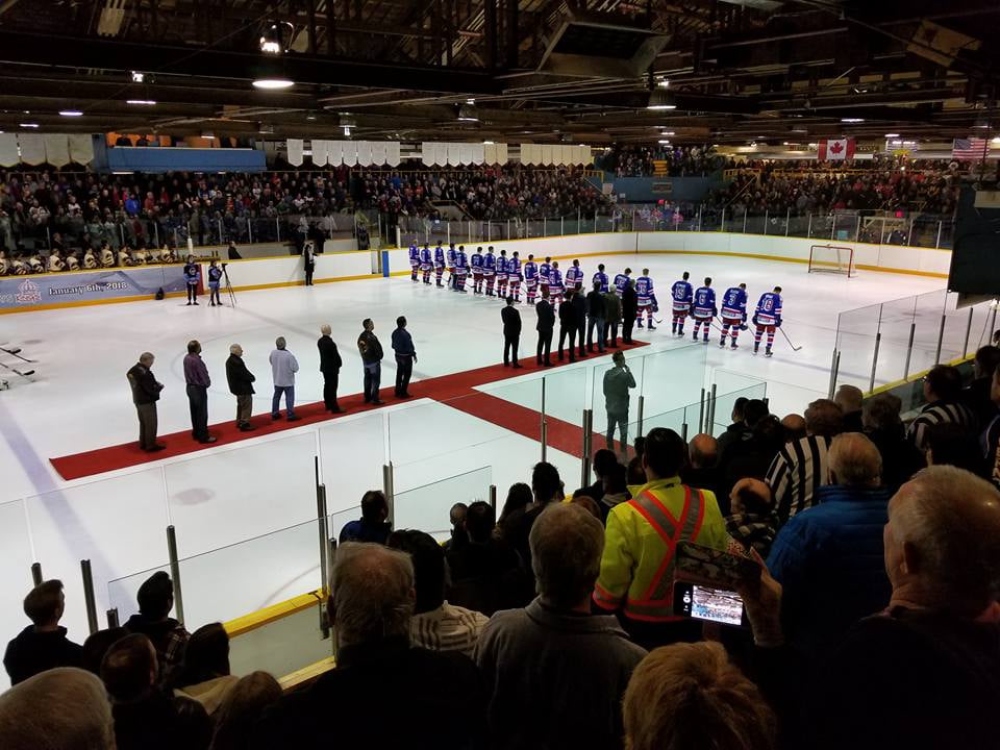 Spectators, hockey players, and dignitaries positioned on a red carpet stand facing a Canadian flag hung at one end of an aging hockey arena.