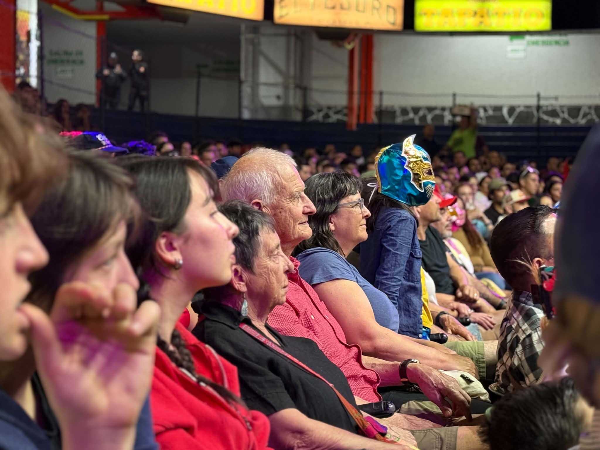 People are seated in an indoor arena facing the right of the frame. One child is wearing a blue and gold lucha libre mask.