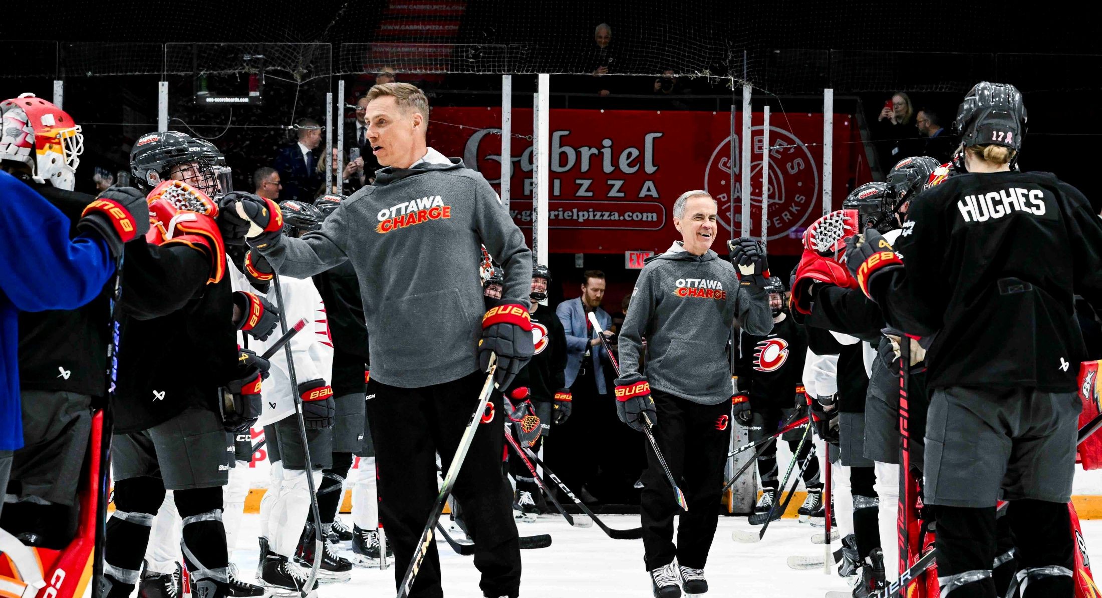 Alexander Stubb, left, and Mark Carney, right, are clad in grey Ottawa Charge hoodies, black pants and black hockey skates. They are arriving on the ice in a hockey arena, greeting players whose backs are turned to the camera and facing them.