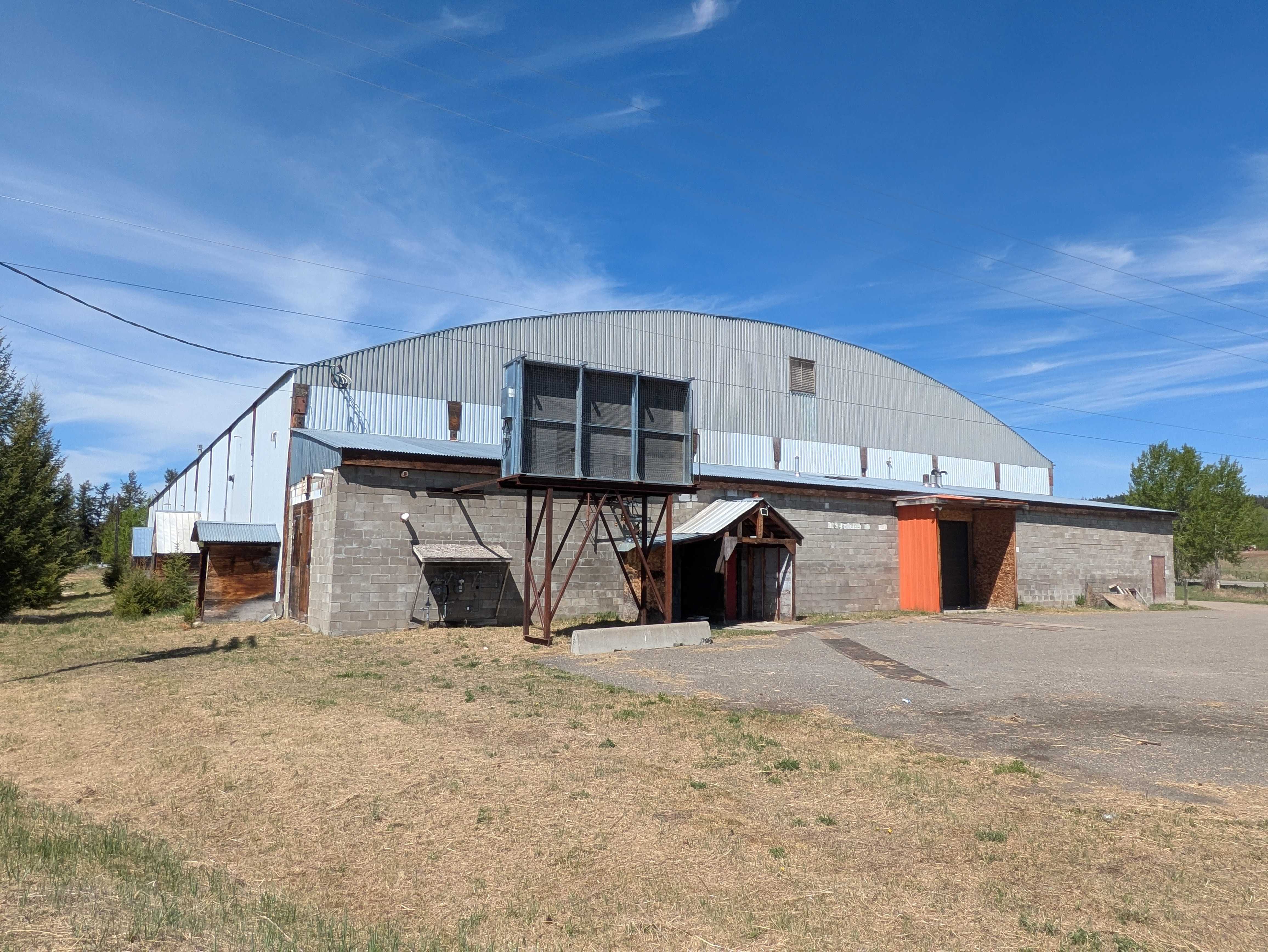 The exterior of the Rolf Zeis Memorial Arena is a low, non-descript grey and light blue building with a driveway to the right of the frame. To the left of the frame is an expanse of dry grass on a bright day.
