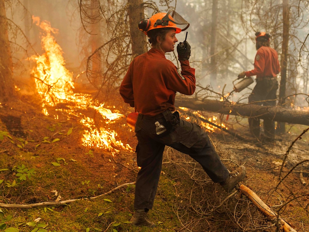 A female wildfire fighter wears a hard hat with a visor and a red uniform shirt while holding up a handheld radio. She's standing in a forest with flames and smoke around her.