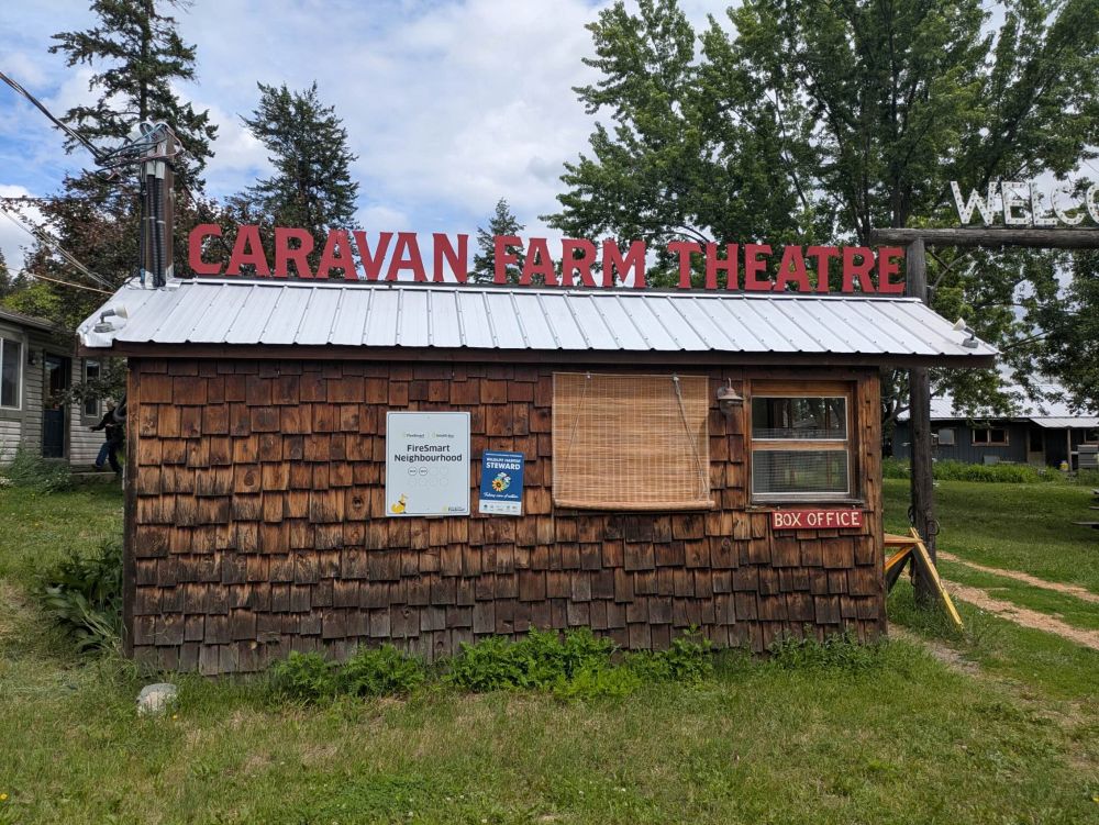 A short building with brown shingled walls and a tin roof bears the words &ldquo;Caravan Farm Theatre&rdquo; in red letters atop its roof. To the right under a window is a hand painted sign that reads &ldquo;box office.&rdquo; The building sits in grass against a stand of trees.