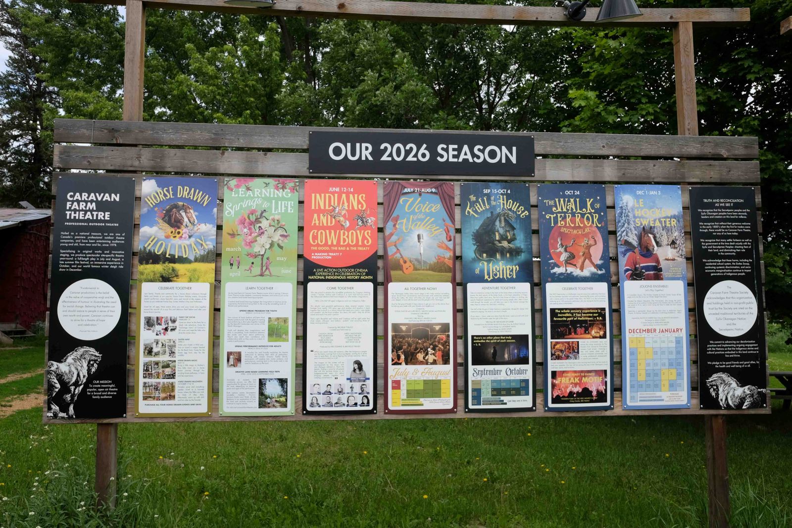 A large, wide outdoor wooden stand displays posters for Caravan Farm Theatre&rsquo;s 2026 season. In the background is a stand of trees.