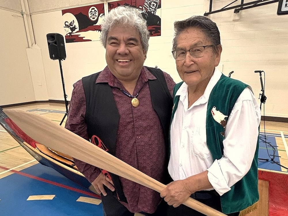 Two Indigenous men wearing collared shirts and vests stand beside each other in front of a canoe in a gymnasium. One holds a canoe paddle.