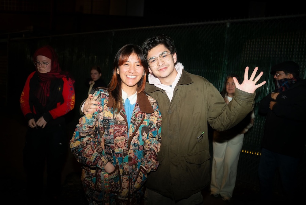 Two young people stand together waving in a dark indoor venue space. The woman on the left has long dyed burgundy hair and she is wearing a patterned jacket. The man on the right has short dark curly hair, glasses and an olive green jacket. He is waving. 