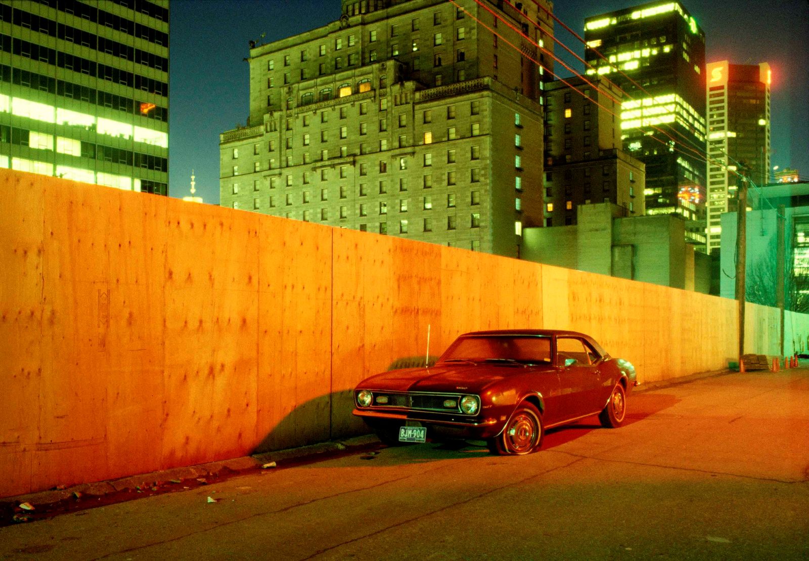 A 1981 colour photograph of a burgundy Camaro in downtown Vancouver alley. It is parked against a wall of plywood, behind which tall towers stand at night.