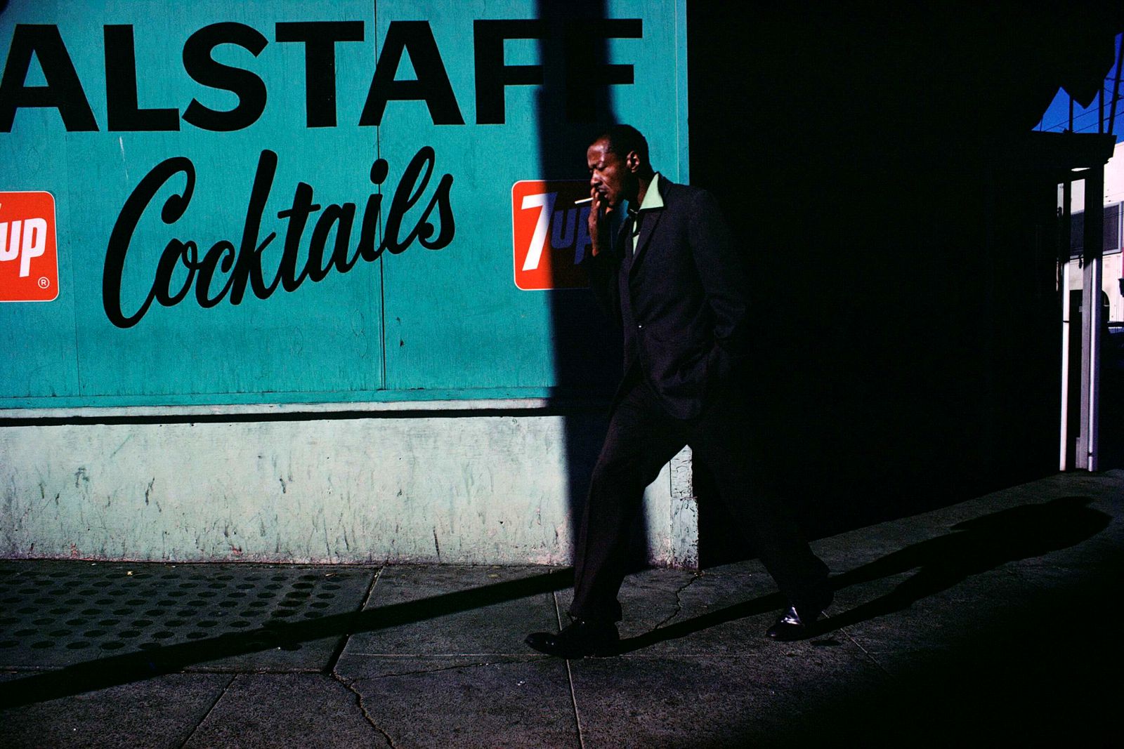 A man in a suit stands in shadows against a post to the right of the frame. He is smoking a cigarette against a teal wall painted with a sign advertising cocktails.