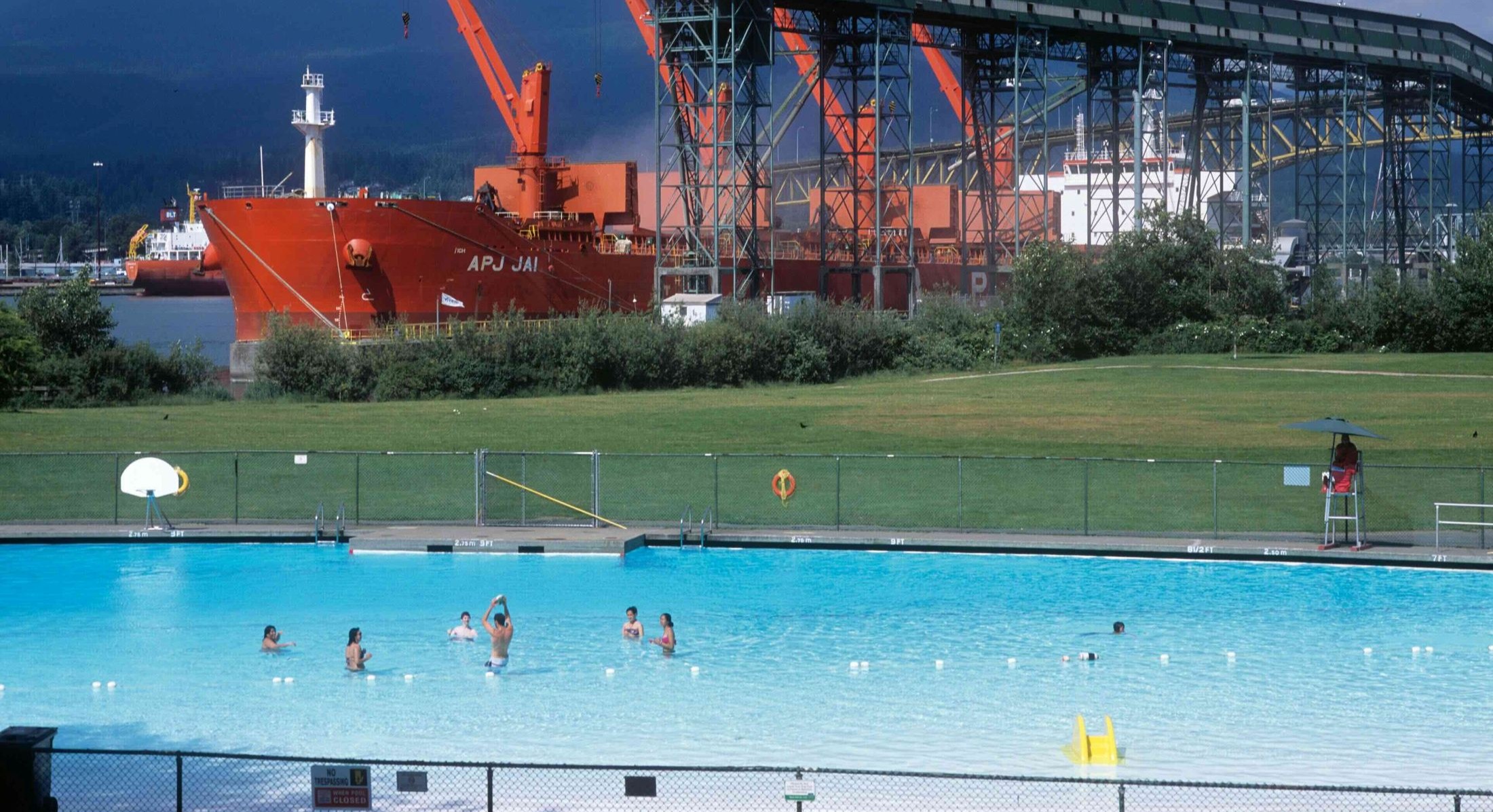 A panorama photo of New Brighton Park in Vancouver features an outdoor public pool with people playing a game with a ball in the water. Behind them is an industrial scene; a large boat on the docks and the Ironworkers Memorial Bridge.