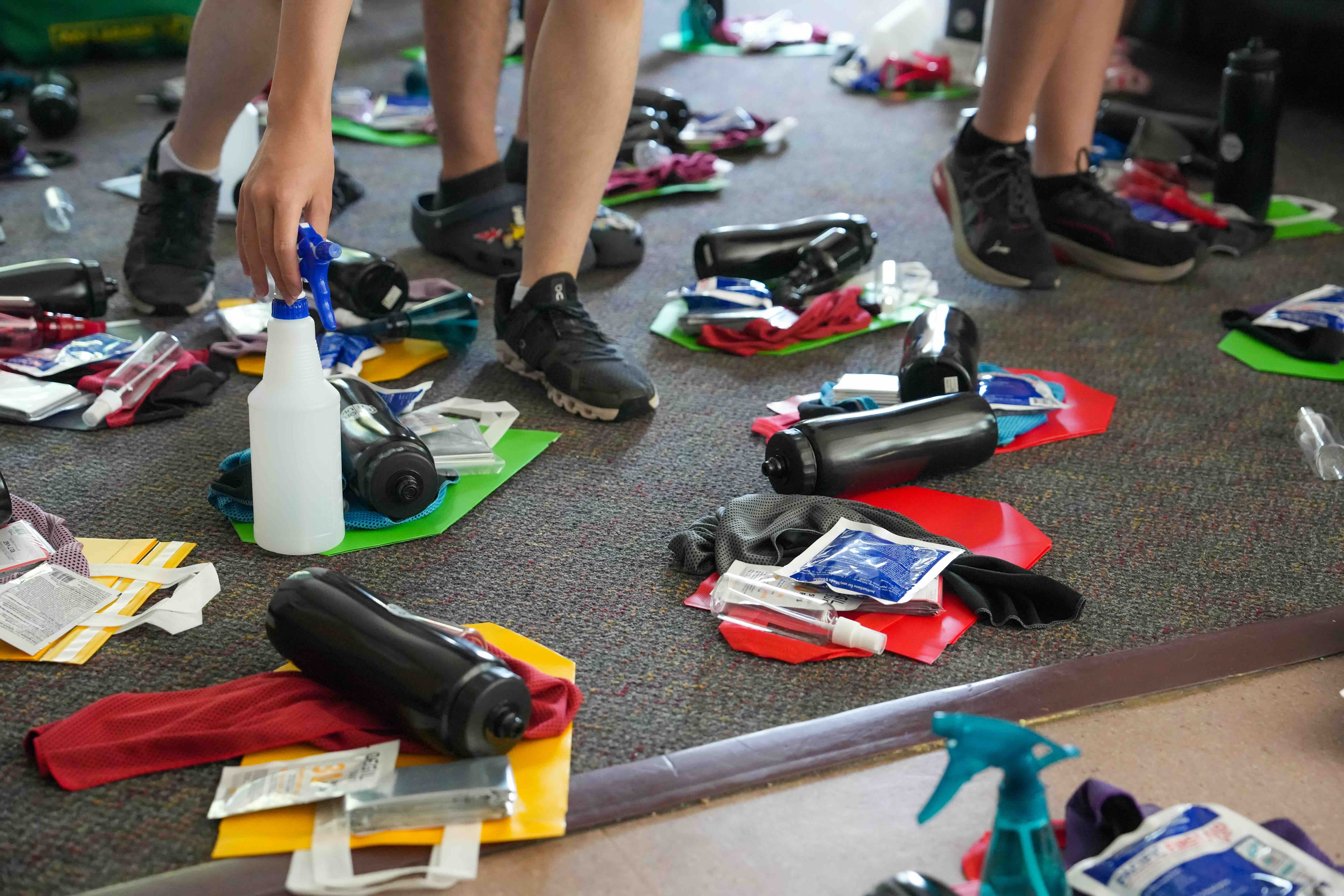 Supplies are laid out in neat bundles across a grey carpet. Bare legs with black sneakers and Crocs step among them.