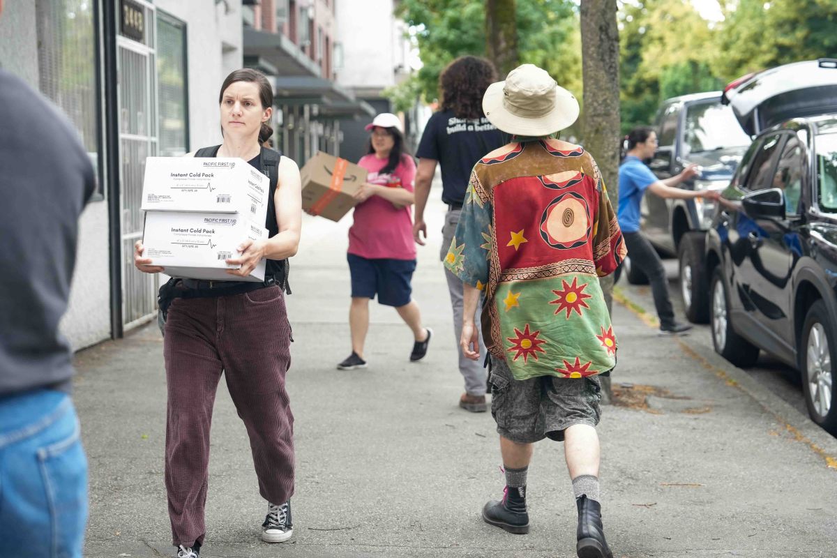 People walk down a sidewalk carrying boxes; some are walking with their backs to the camera towards vehicles parked to the right of the frame to help unload them.