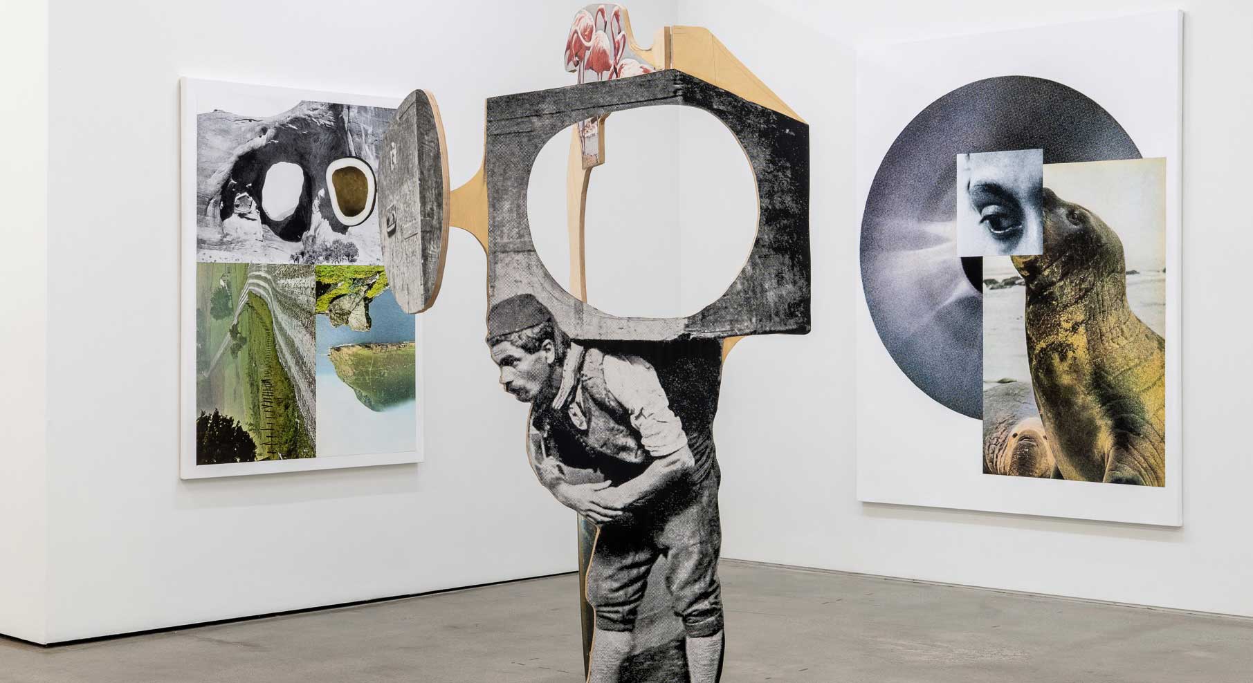 A sculpture in the centre of the frame depicts a black-and-white photo of a man in a cap and old-fashioned clothing carrying a large, abstract shape on his back. In the background are large pieces of multimedia artwork on white walls.