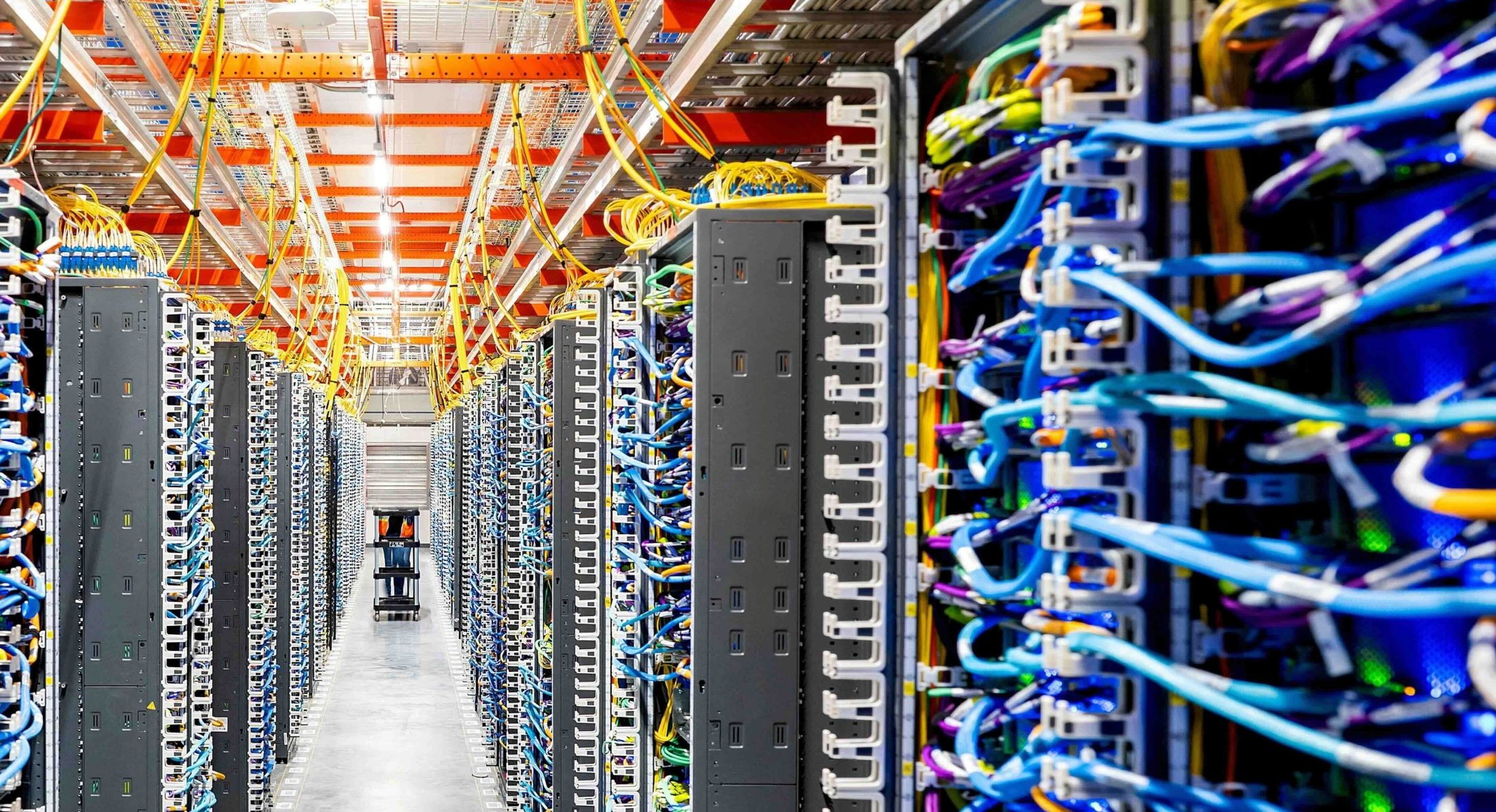 A colourful network of wires in the hallways of an AI data centre. There are blue and purple wires in banks on the floor and yellow and orange ones on the ceiling.