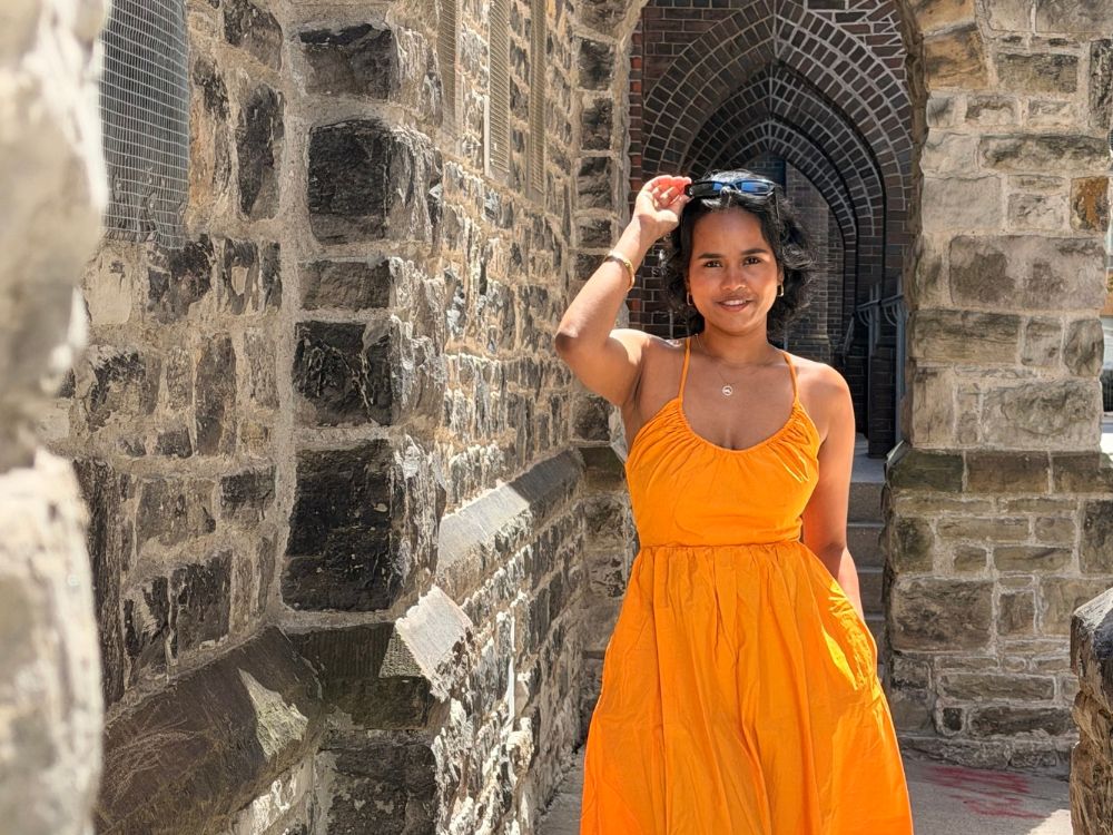 Shubhalaxmi Patil is standing outdoors in an area with brick walls. She holds her sunglasses on her head and looks directly at the camera. She is wearing a yellow dress.