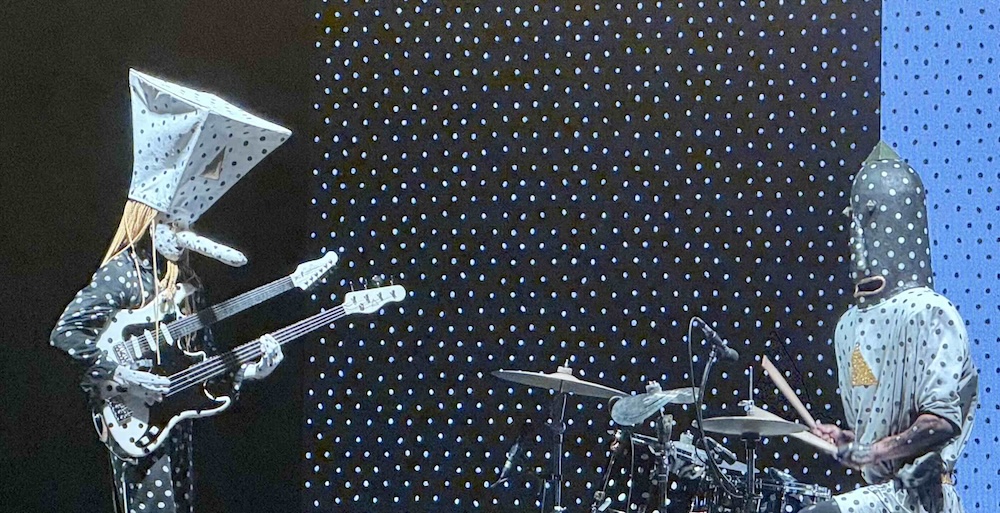 Two people in abstract black and white dotted costumes and masks play guitar (left) and drums (right) onstage. The stage backdrop features a black and white dot pattern similar to the fabric of their costumes.